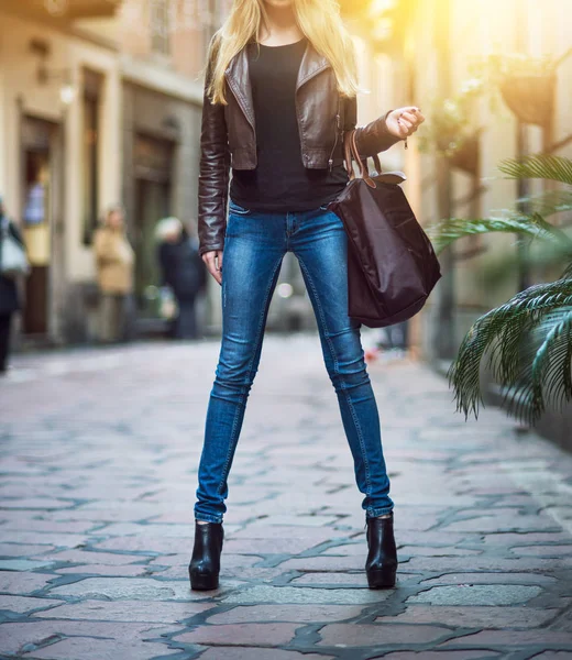 Fashionable young blonde girl with long legs wearing blue jeans, leather brown coat and holding a bag walking and shopping on city street in old town — Stock Photo, Image