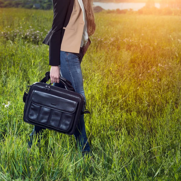 Student girl walking and carrying black leather laptop case bag outdoors on sunny green grass meadow.