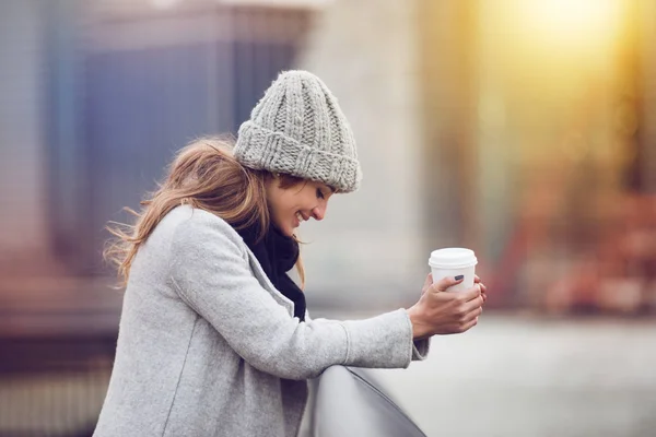 Счастливая молодая женщина пьет кофе рядом с небом Нью-Йорка в зимней одежде и улыбается. Красивая женщина держит чашку кофе на открытом воздухе в городе . — стоковое фото