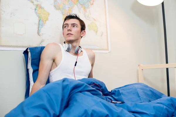 Молодой человек проснулся и слушал музыку в наушниках в постели дома. Красивый студент сидит в постели в белой футболке . — стоковое фото