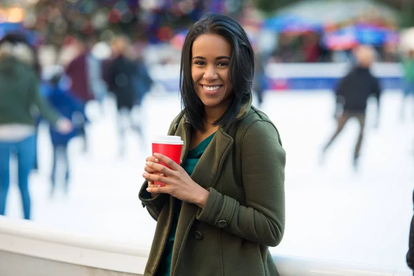 Beautiful happy smiling latin woman drinking coffee at city ice rink in front of Christmas tree. Happy young adult woman enjoying Christmas time in the city and drinking coffee and looking at camera. — Stock Photo, Image