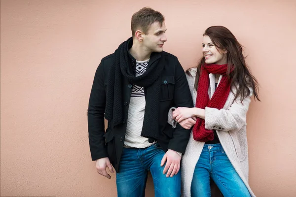 Happy young fashionable couple in love wearing warm stylish winter outfits with jacket, jeans, sweater and scarf standing near the wall and looking on each other smiling. — Stock Photo, Image