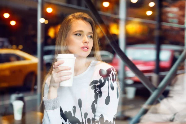 Bella donna che beve caffè in caffetteria dalla tazza di caffè di carta bianca e guardando la strada della città attraverso la finestra. Ragazza che tiene in mano la tazza di caffè . — Foto Stock