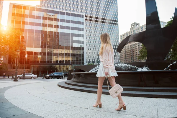 Hermosa joven con piernas largas caminando por la calle de la ciudad vistiendo falda corta y camiseta rosa y sosteniendo una bolsa. Chica vistiendo ropa de verano estilo calle . — Foto de Stock