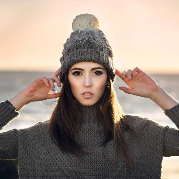 Beautiful young woman wearing grey knitted hat and warm knitted sweater posing outdoors and looking in camera — Stock Photo, Image