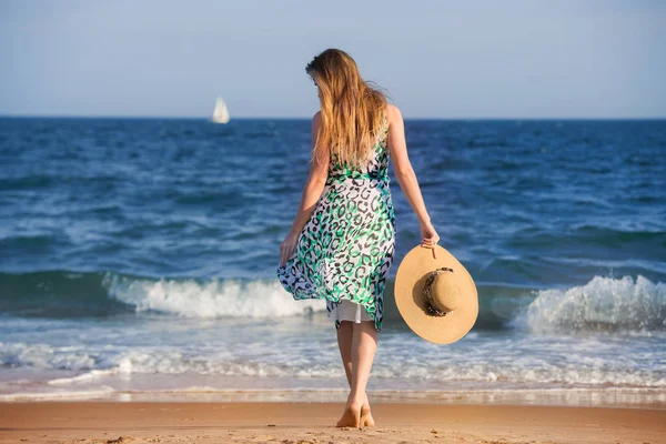 Young barefoot woman with hat walking on ocean beach at sunny hot day and enjoy the vacation. Girl wearing summer dress walking on sand. — Stock Photo, Image