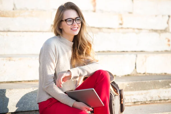 Happy beautiful student girl wearing eyeglasses sitting on college steps in the campus with a bag and tablet — Stock Photo, Image