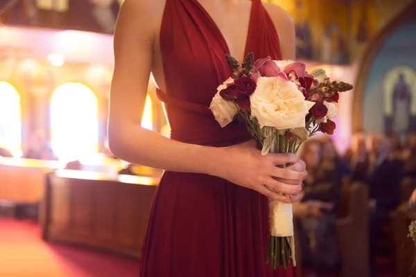 Esküvői ceremónia napja. koszorúslány lány visel elegáns piros ruha gazdaság virág csokor, az esküvői szertartás a katolikus egyház. — Stock Fotó