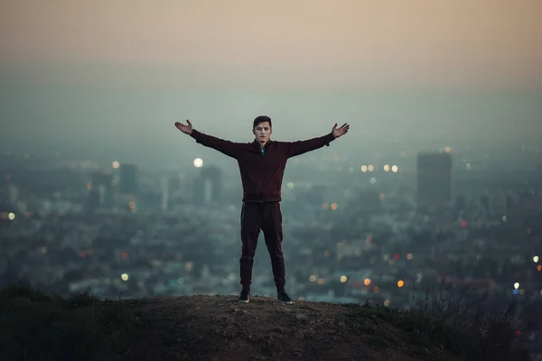 Homem turístico em pé na colina com as mãos levantadas ao lado da grande luz da cidade . — Fotografia de Stock