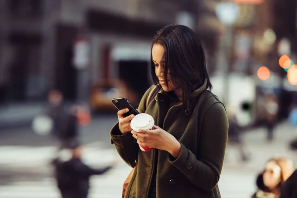 Young adult energetic woman texting on cell phone, drinking coffee and walking to work on city street — Stock Photo, Image