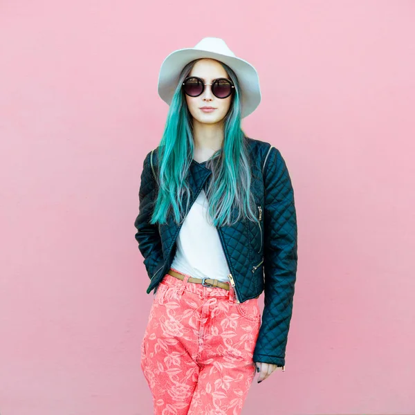 Mulher blogueira jovem e elegante com cabelo azul vestindo roupas de estilo casual com jaqueta preta, chapéu branco, jeans rosa e óculos de sol posando perto da parede . — Fotografia de Stock