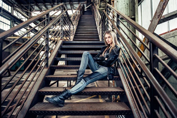 Hermosa mujer posando con traje casual con chaqueta de cuero, zapatos negros y vaqueros de moda. Chica posando en planta industrial . —  Fotos de Stock