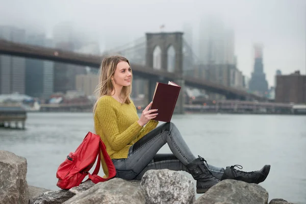 Молодий студент красива дівчина читання книги, сидячи біля міста Нью-Йорк skyline. Навчатися в США університет концепції. — стокове фото