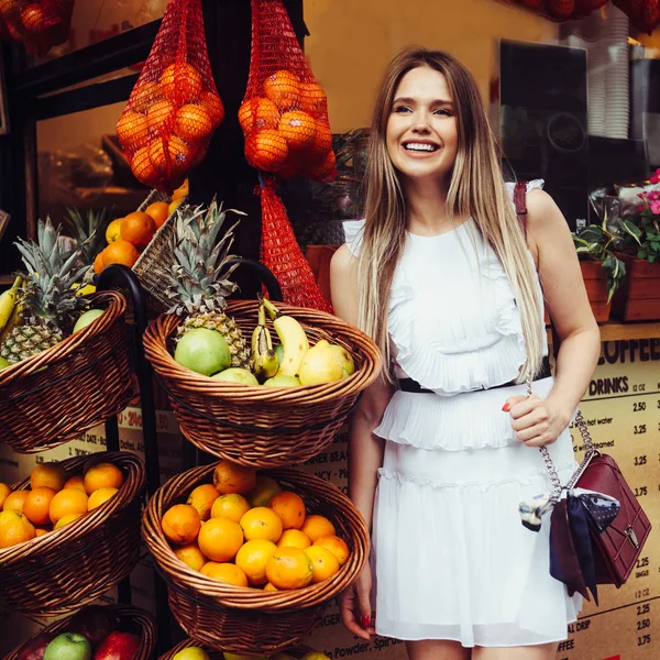 Schöne lächelnde junge erwachsene Frau, die in der Nähe von Streuobstladen mit tropischen Früchten in Körben steht — Stockfoto