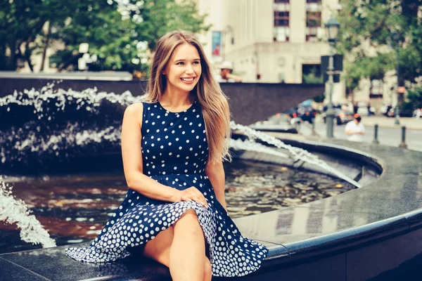 Красива молода усміхнена жінка сидить на фонтані міського парку в літній день одягнена в сукню і дивиться в бік — стокове фото
