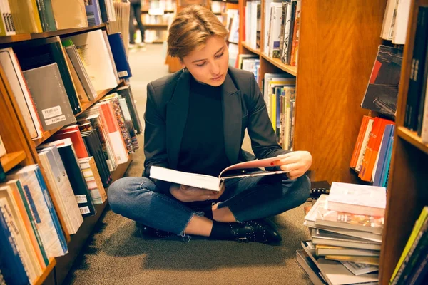 Estudiante sentada en el suelo y leyendo un libro en la biblioteca —  Fotos de Stock