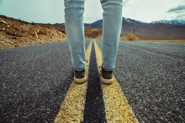 Men`s feet standing on aslphalt desert road with yellow marking lines. Man wearing sneakers and jeans.