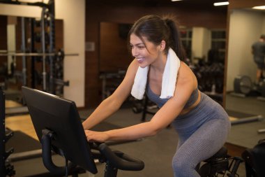 Gülümseyen genç ve güzel bir kadın spor salonunda sabit bir bisiklete biniyor.