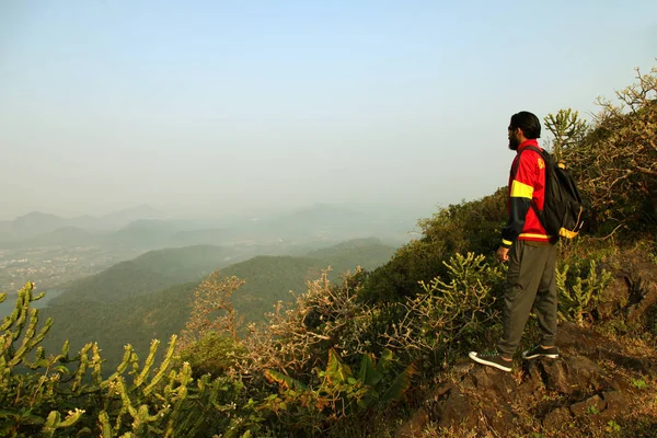 Jovem com mochila e capacete em pé no topo de uma montanha e desfrutando de vista para o vale — Fotografia de Stock