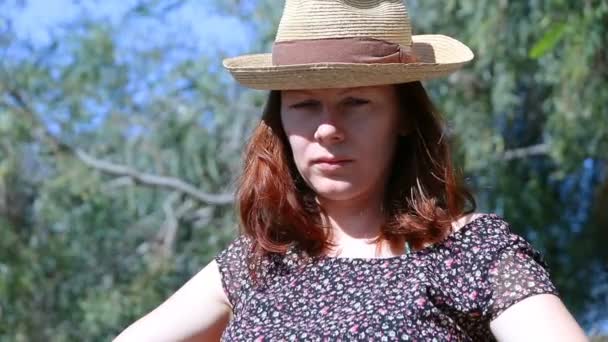 Young beautiful woman with red hairs and a straw hat sitting outside looking at the camera and then looking up seriously — Stock Video