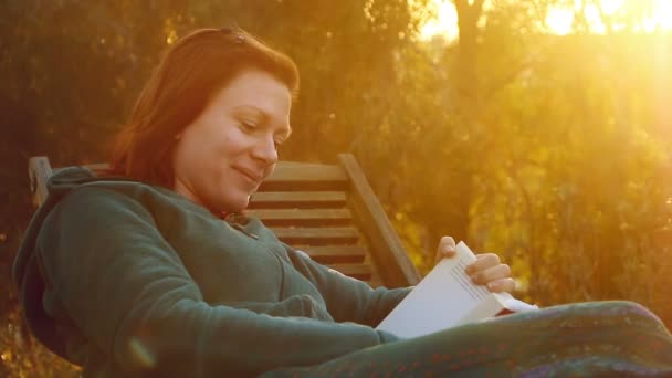 Giovane bella donna sorridente si rilassa su una sedia a sdraio leggendo un libro in retroilluminazione tramonto — Video Stock