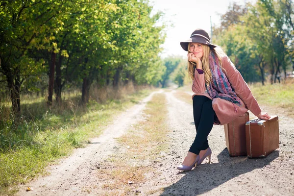 スーツケース 日当たりの良い屋外の背景と帽子エレガントなおしゃれなコートを着ている美しい若い女性 — ストック写真