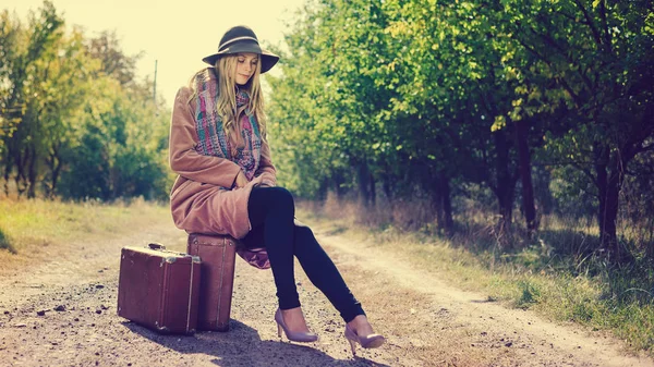 スーツケース、屋外の日当たりの良い背景を持つ帽子エレガントなおしゃれなコートを着ている美しい若い女性 — ストック写真