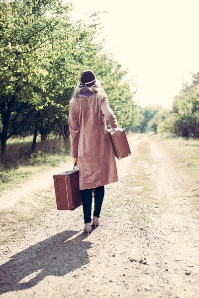 Back view of female wearing hat coat walking away on rural road. She carrying old suitcase over sunny outdoors background
