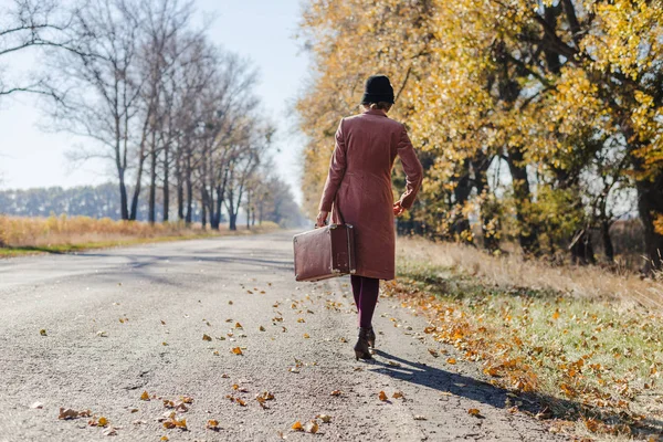 ピンクのヴィンテージコートを着た若い赤毛の女性と黄金の黄色の秋の木と公園の道路に沿って歩いてレトロなスタイルのスーツケース付きの帽子。屋外の秋の庭のリラクゼーション、旅行のコンセプト — ストック写真