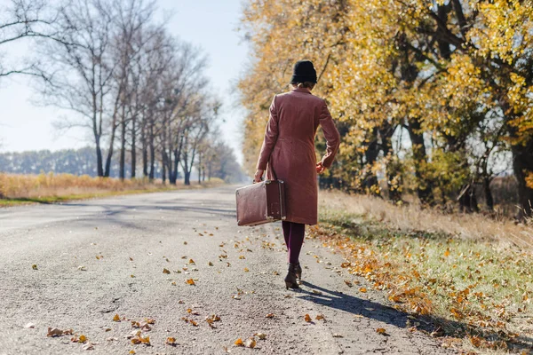 Mujer pelirroja joven en abrigo vintage rosa y sombrero con maleta de estilo retro que se aleja a lo largo de un camino del parque con árboles otoñales de color amarillo dorado. Relajación jardín otoño al aire libre, concepto de viaje —  Fotos de Stock