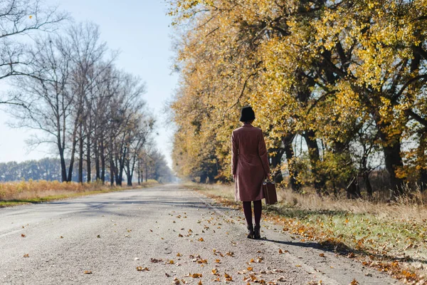 Giovane donna rossa in cappotto rosa vintage e cappello con valigia in stile retrò che si allontana lungo una strada del parco con alberi autunnali giallo oro. Outdoor autunno giardino relax, concetto di viaggio — Foto Stock