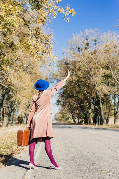 Mulher jovem ruiva senhora em casaco vintage rosa e chapéu com mala em estilo retro caminhando ao longo de uma estrada do parque com árvores outonais amarelas douradas. Outono exterior jardim relaxamento, conceito de viagem — Fotografia de Stock