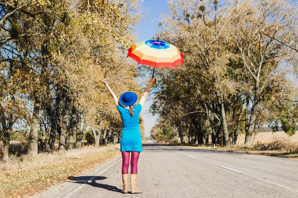 Giovane donna in stivali di gomma, con ombrello colorato e valigia d'epoca autostop su strada d'autunno vuota — Foto Stock