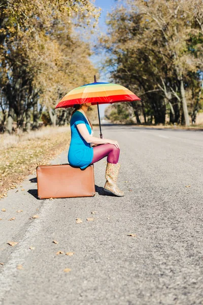 Kobieta z kolorowym parasolem i zabytkową walizką siedzi na pustej drodze — Zdjęcie stockowe