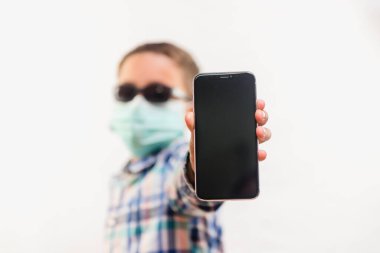Tek kullanımlık maske takan ve beyaz arka plan üzerinde cep telefonu tutan bir çocuk.