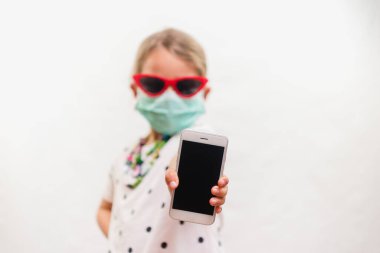 Tek kullanımlık maske takan küçük sarışın kız ve beyaz arka planda cep telefonu tutan güneş gözlüğü.