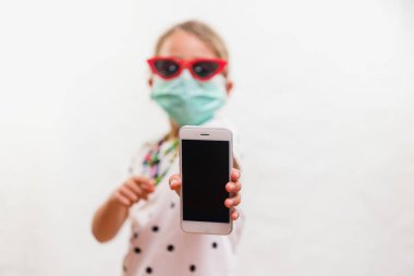 Tek kullanımlık maske takan küçük sarışın kız ve beyaz arka planda cep telefonu tutan güneş gözlüğü.