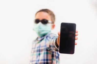 Tek kullanımlık maske takan ve beyaz arka plan üzerinde cep telefonu tutan bir çocuk.
