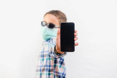 Tek kullanımlık maske takan ve beyaz arka plan üzerinde cep telefonu tutan bir çocuk.