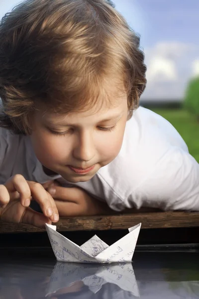 Menino feliz jogando com um origami navio de papel na lagoa solar ri — Fotografia de Stock