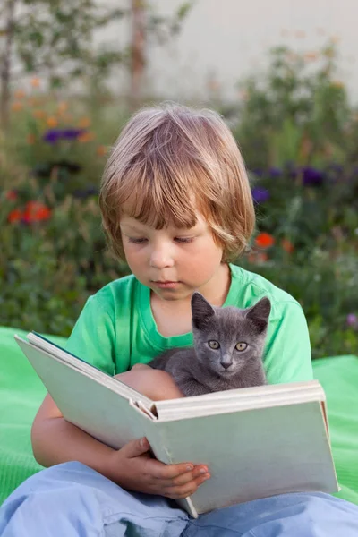 Мальчик читает книгу с котенком во дворе, ребенок с чтением домашних животных — стоковое фото