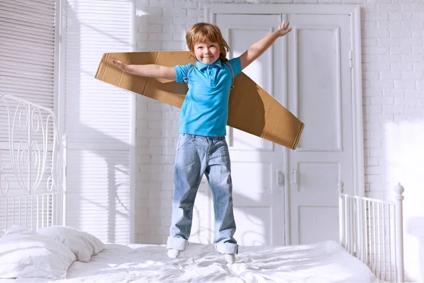 Kind met kartonnen vleugels springen op bed in de slaapkamer — Stockfoto
