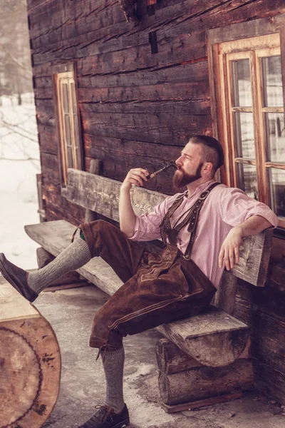 Young Bavarian Man Leather Pants Pipe Hand Sits Wooden Bench — Stock Photo, Image