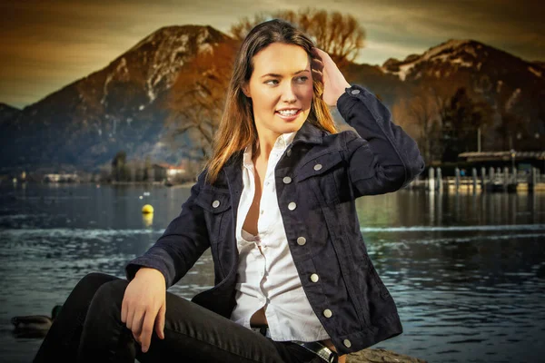 女人穿着斜纹棉布夹克和牛仔裤坐在湖边 — 图库照片