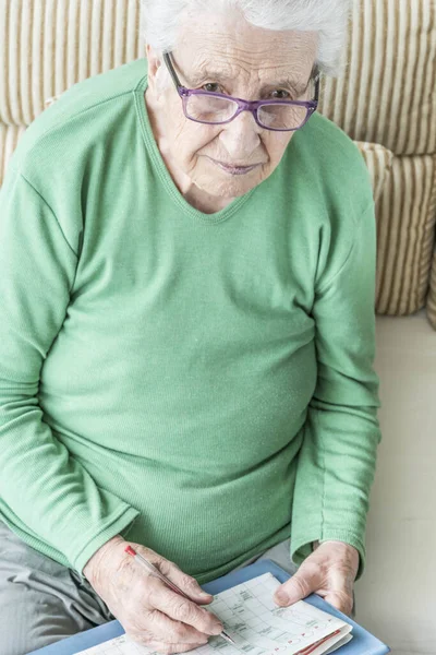 Evde Bulmaca Çözen Yaşlı Bir Kadın Kameraya Bakıyor — Stok fotoğraf