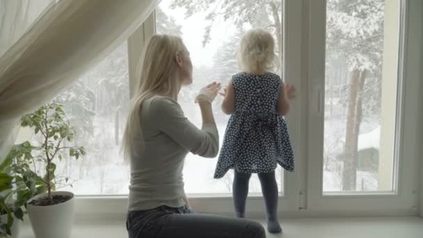Madre está mostrando el paisaje invernal a su pequeña hija a través de una ventana — Vídeos de Stock
