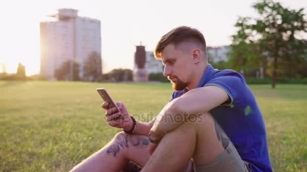 Hombre sentado en la hierba y y voltear el teléfono inteligente — Vídeo de stock