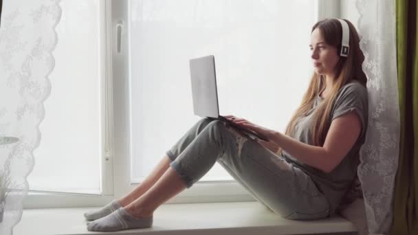 Женщина с наушниками сидит на подоконнике и использует ноутбук — стоковое видео