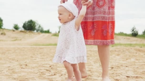 Feliz bebé está dando los primeros pasos con la madre en la playa de arena — Vídeos de Stock