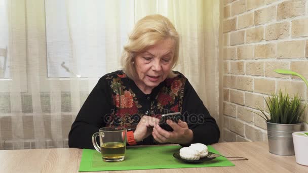 Ευτυχισμένος ηλικιωμένη γυναίκα πίνοντας τσάι και χρησιμοποιώντας ένα smartphone — Αρχείο Βίντεο